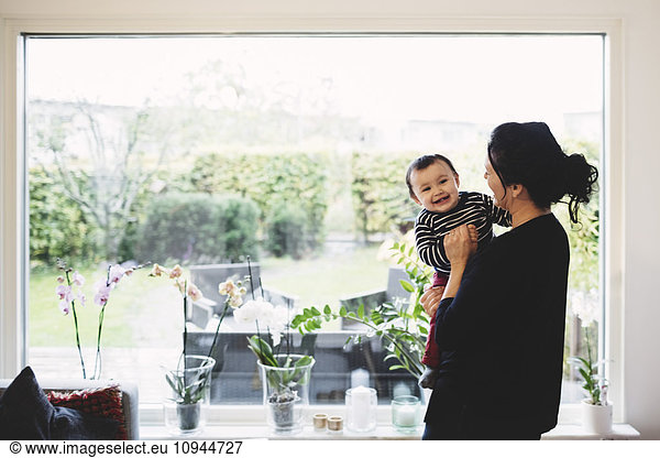 Mother carrying smiling baby girl while standing by window at home