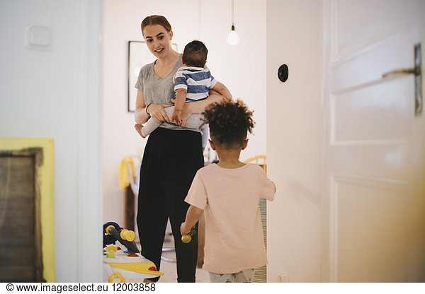 Mother carrying toddler looking at son while standing in domestic room