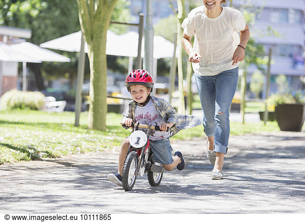 Mother chasing son riding bicycle with helmet in sunny park