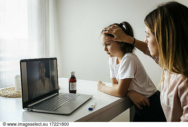 Mother checking daughter while attending video call with doctor over laptop at home