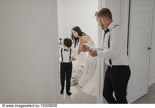 Mother dressing son for wedding while father standing in doorway at home
