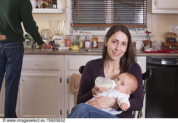 Mother feeding her son from bottle and father cleaning kitchen