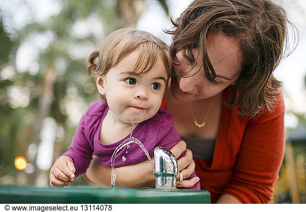 Mother helping cute daughter in drinking water from fountain
