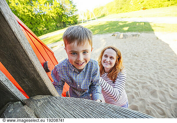 Mother helping son to climb outdoor play equipment at playground