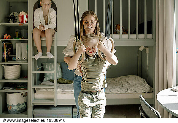 Mother helping son with down syndrome swinging on gymnastics rings in bedroom