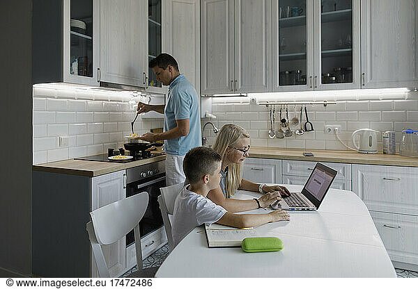 Mother helping son with homework on laptop at table by man cooking in kitchen