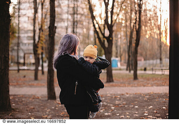 Mother holding cute baby son in park during autumn at sunset