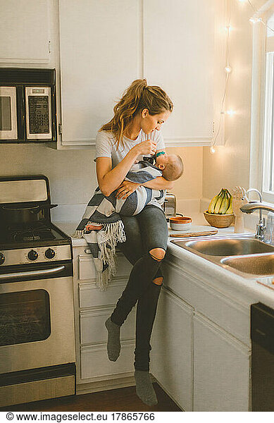 Mother holding infant while sitting on the counter in small kitc