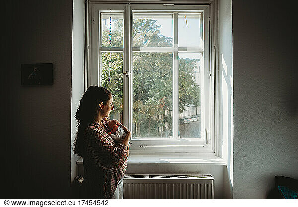 Mother holding newborn baby in room looking out the window