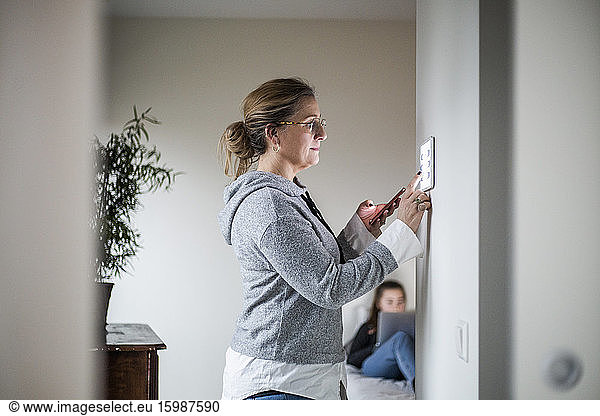 Mother holding smart phone while using digital tablet on wall with daughter in background at smart home