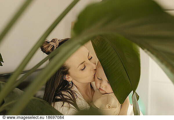Mother kissing daughter seen through monstera plant at home