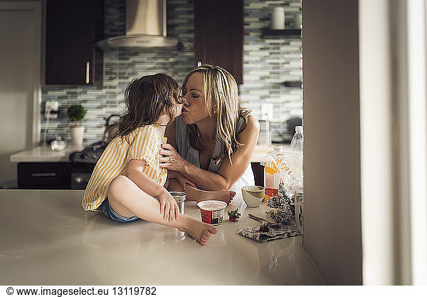 Mother kissing daughter sitting on kitchen counter at home