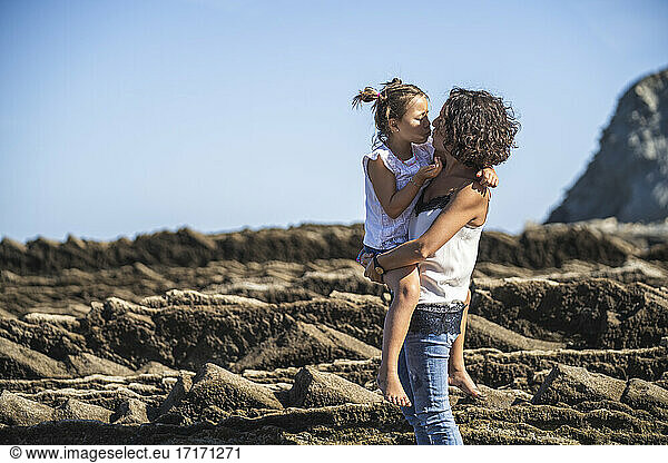 Mother kissing daughter while standing on flysch against blue sky