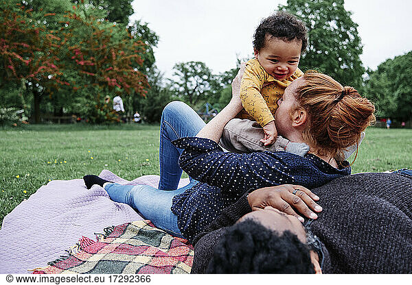 Mother kissing son while leaning on man resting on picnic blanket at park