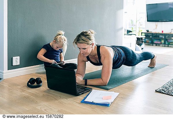 Mother looking at laptop and doing planks while daughter using smart phone in living room