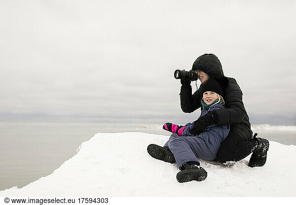 Mother photographing through camera sitting with daughter on snow in winter