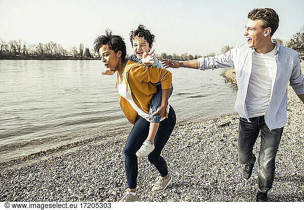 Mother piggybacking cheerful boy while running by lakeshore