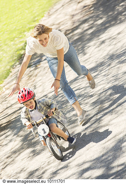 Mother pushing son with helmet on bicycle in sunny park