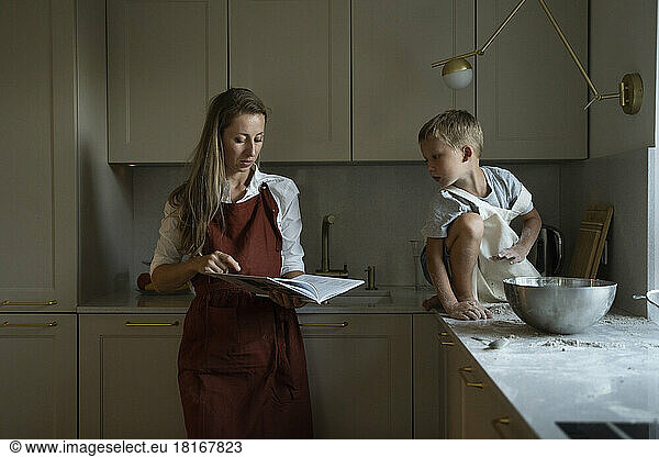 Mother reading recipe book with son sitting on kitchen counter