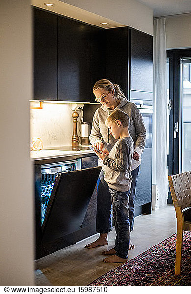Mother teaching son to use mobile app while operating dishwasher in kitchen at home