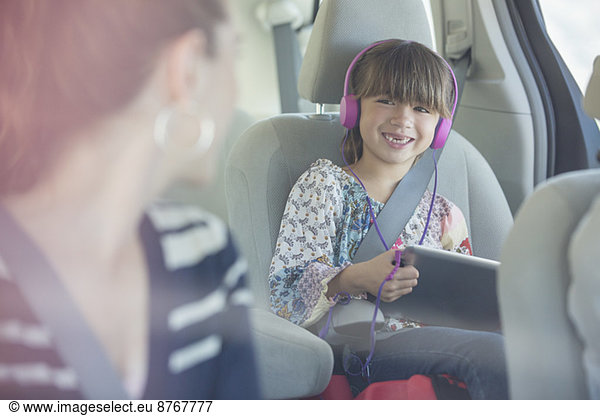 Mother turning and smiling at daughter with headphones and digital tablet in back seat of car