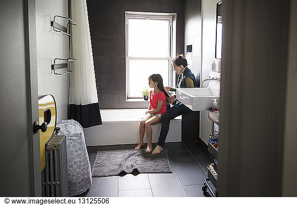 Mother tying hair of daughter while sitting in bathroom