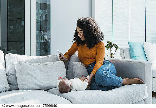 Mother using smart phone sitting by baby boy on sofa at home