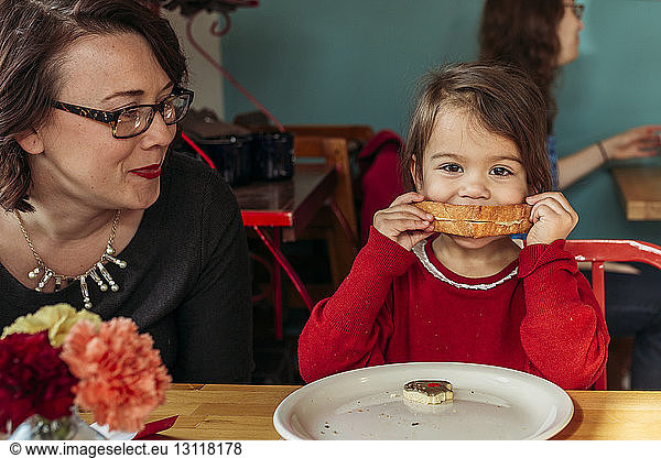 Mother watching daughter eating bread at restaurant table