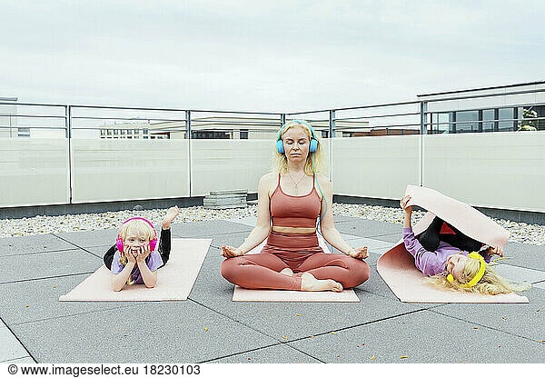 Mother wearing headphones practicing yoga with daughters at rooftop