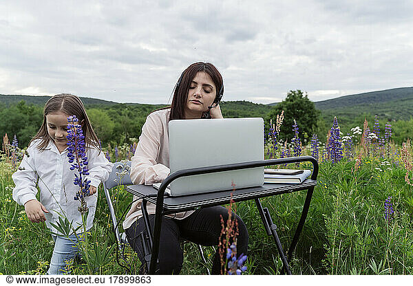Mother with eyes closed at desk by daughter playing in meadow