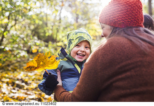 Mother with happy boy holding leaf in forest during autumn