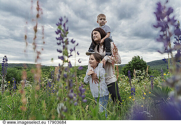 Mother with son on shoulders by daughter in lupine flowers meadow