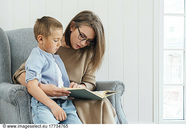 Mother with son reading book sitting on armchair at home