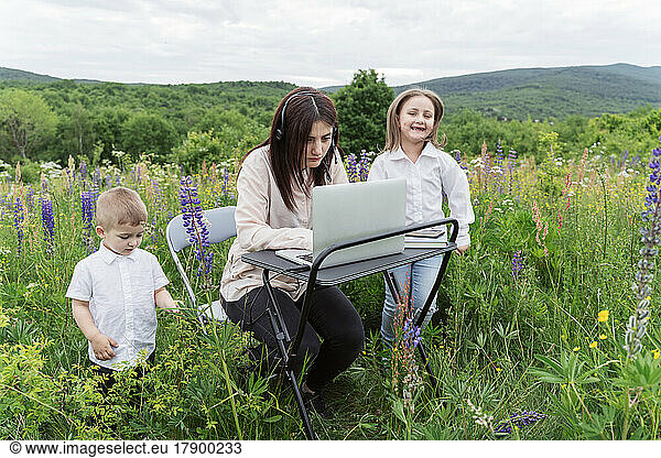 Mother working on laptop by playful children in meadow