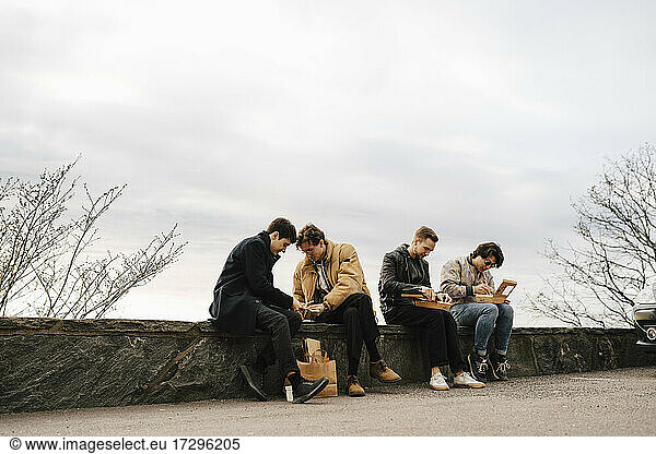 Multi-ethnic male friends having food while sitting on retaining wall against sky
