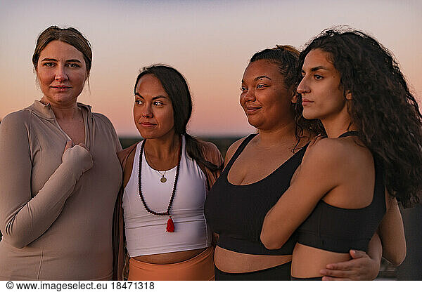 Multiracial female friends standing with arms around at sunset