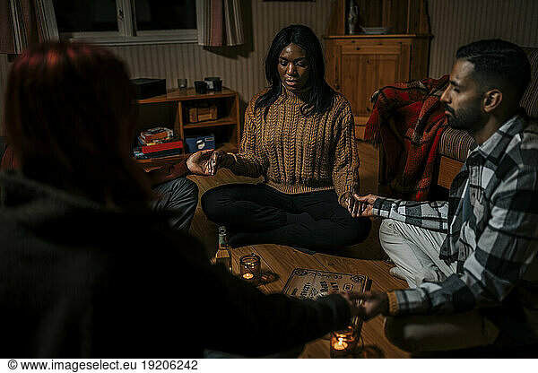 Multiracial friends holding hands sitting with ouija board in log cabin