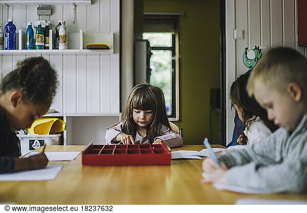 Multiracial male and female students writing on paper at table in kindergarten