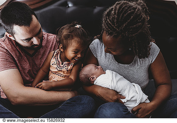 Multiracial parents holding smiling 2 yr old girl and swaddled baby