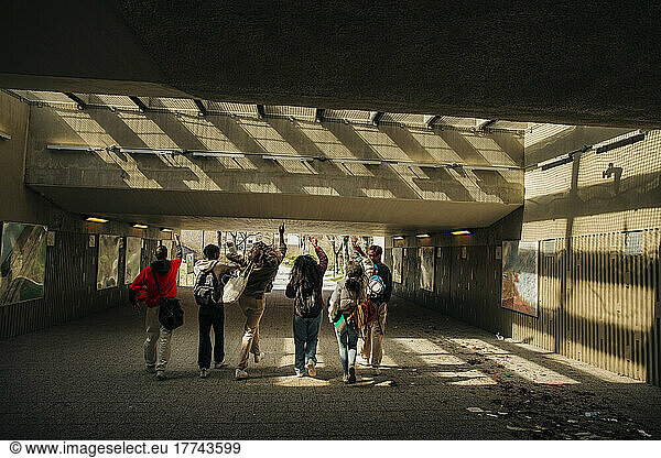 Multiracial young friends with hands raised walking together through underpass