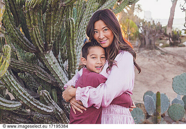 Mutter  die ihren Sohn vor einem großen Kaktus umarmt  beide schauen in die Kamera.
