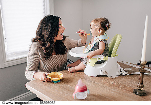 Mutter füttert Baby-Tochter im Kindersitz auf dem Küchentisch