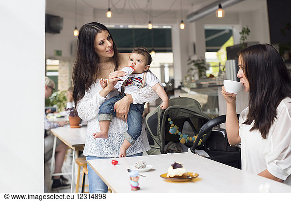 Mutter hält ihren kleinen Sohn (6-11 Monate) und unterhält sich mit einem Freund in einem Café