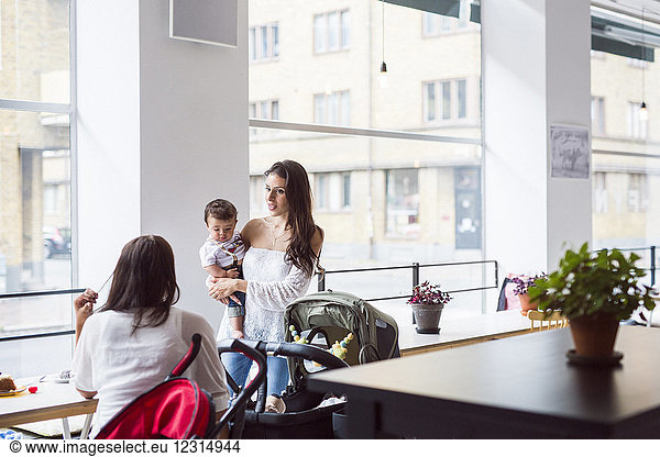 Mutter hält ihren kleinen Sohn (6-11 Monate) und unterhält sich mit einem Freund in einem Café