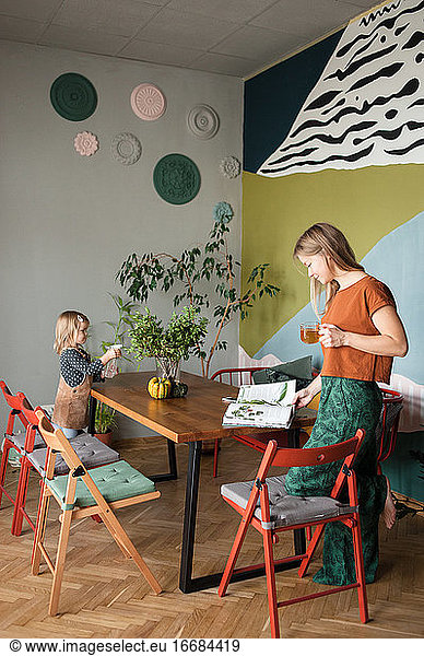 Mutter liest am Tisch stehend eine Zeitschrift  Kind gießt Pflanzen auf dem Tisch