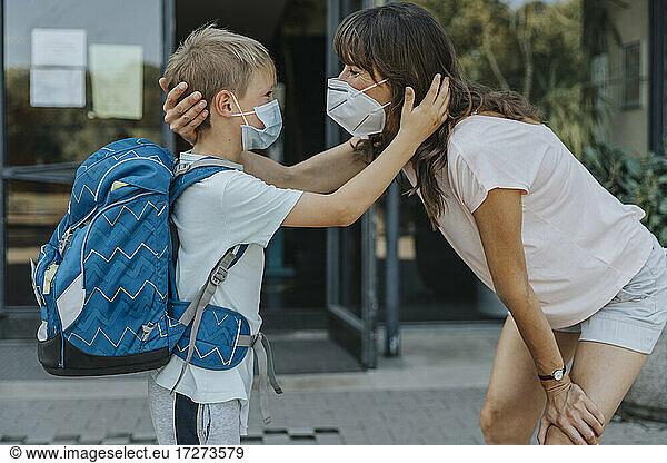Mutter und Sohn mit Schutzmaske stehen sich vor einem Schulgebäude Auge in Auge gegenüber