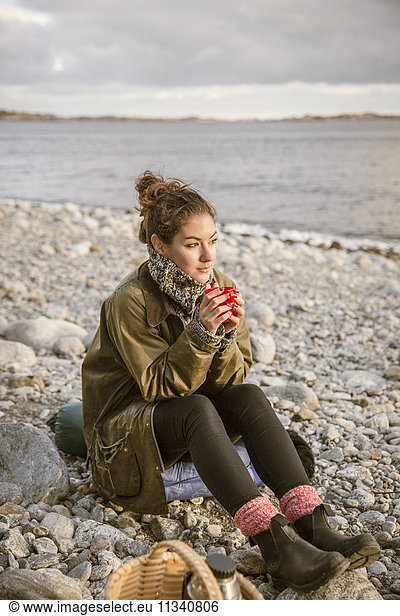 Nachdenkliche Frau sitzt auf einem Felsen und trinkt am Strand.