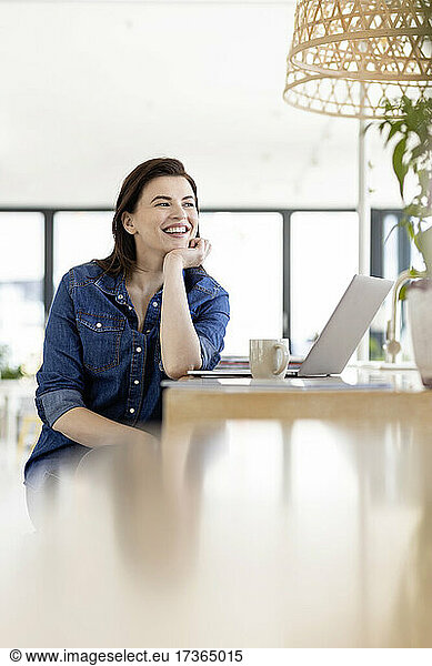 Nachdenkliche Geschäftsfrau mit Hand am Kinn am Laptop sitzend am Arbeitsplatz