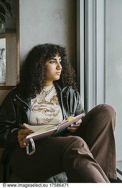 Nachdenkliche junge Frau  die mit einem Buch sitzt und aus dem Fenster der Universität schaut