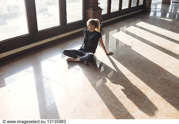 Nachdenkliche junge Frau in voller Länge  die auf dem Fliesenboden sitzt und durch das Fenster schaut